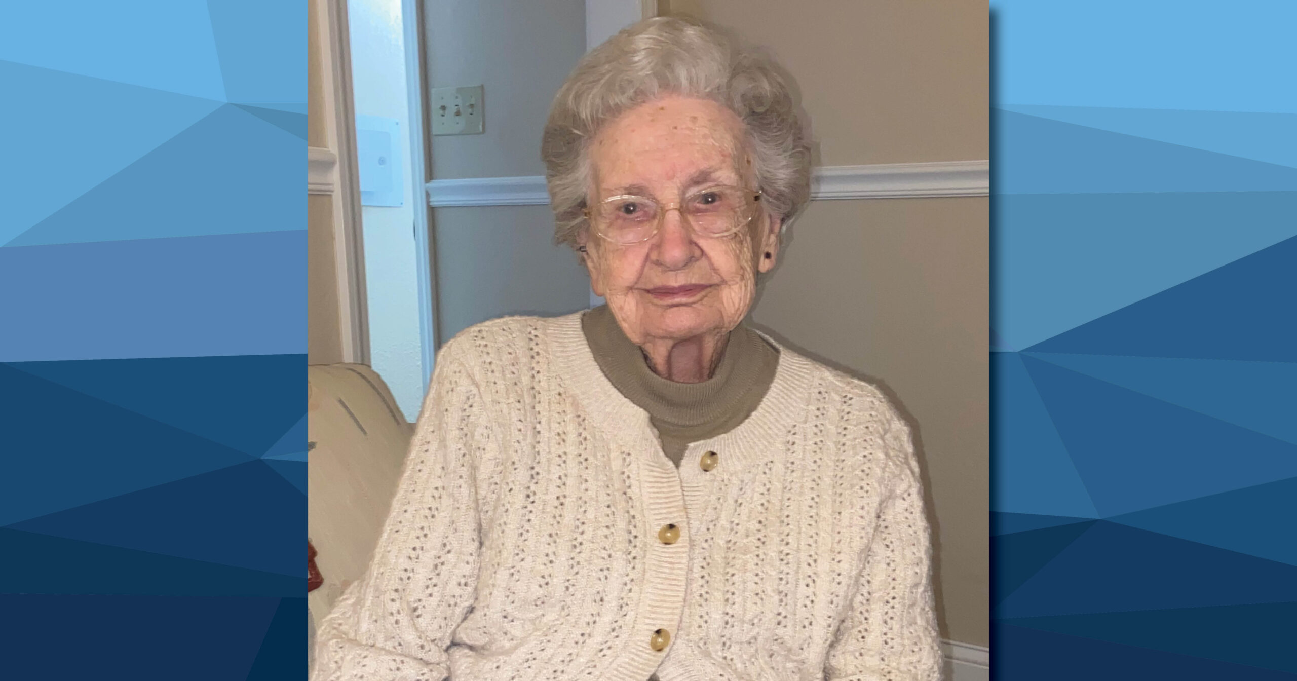 Bernell Hogan sits in a tan chair wearing a cream-colored cardigan and grey shirt. She is wearing glasses and has short grey hair.