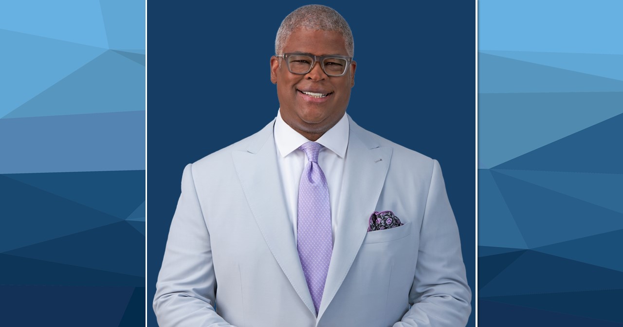 Charles Payne smiles in a grey suit jacket, lavendar tie, and white shirt in front of a navy background.