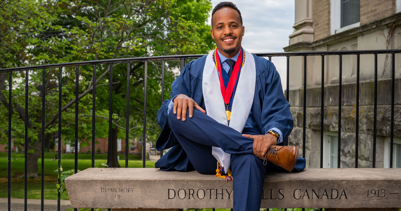 Sayidcali Ahmed smiles and sits on a stone bench in front of Newnam Hall wearing a blue graduation gown, blue pants, brown shoes, a white stole and red ribbon with a gold medallion.