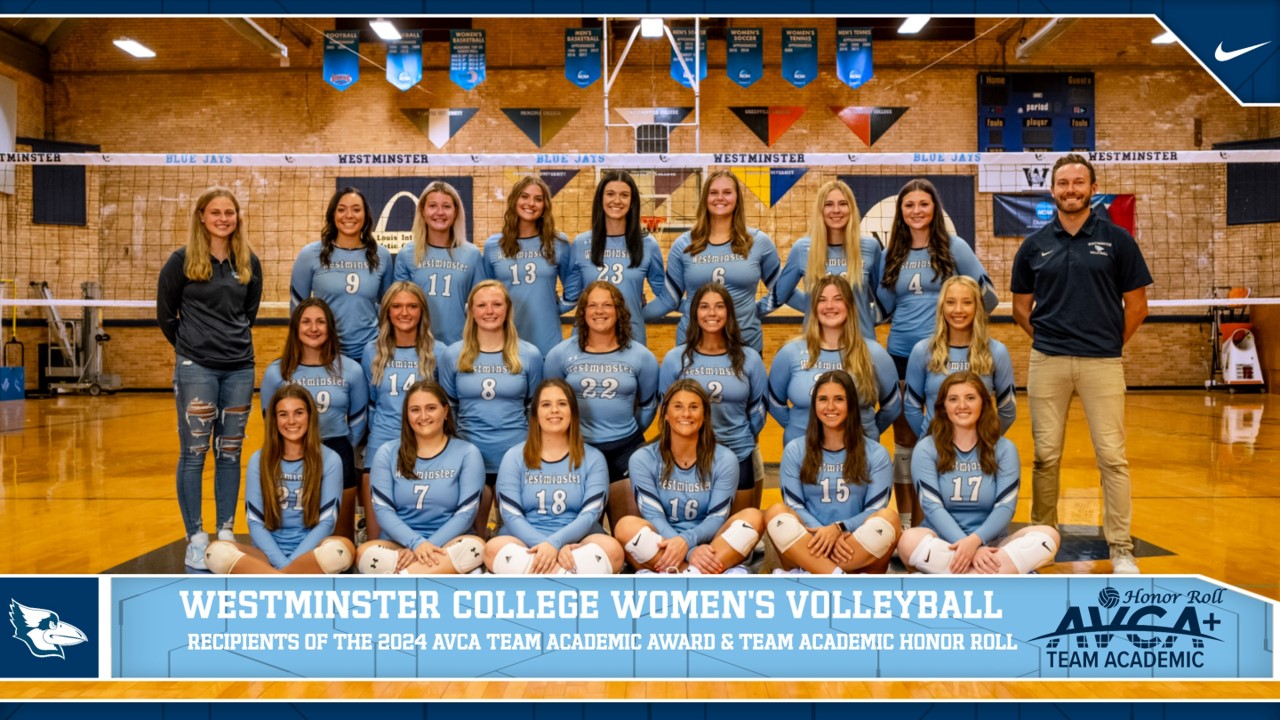 The Blue Jays women's volleyball team poses in light blue uniforms in front of a volleyball net in the Historic Gymnasium, flanked by their coaches on the left and right.