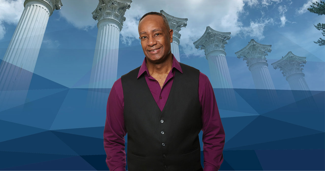 Rich Braham, wearing a magenta shirt and black blazer, stands in front of a blue backdrop with Westminster's Columns in the background.