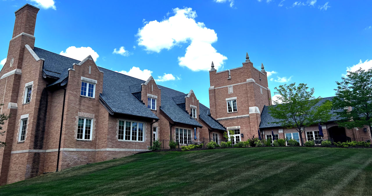 Mueller Leadership Hall, with its light-brown bricks and tall gables, stands proudly on campus amid bright green grass and a blue sky with puffy, white clouds overhead.