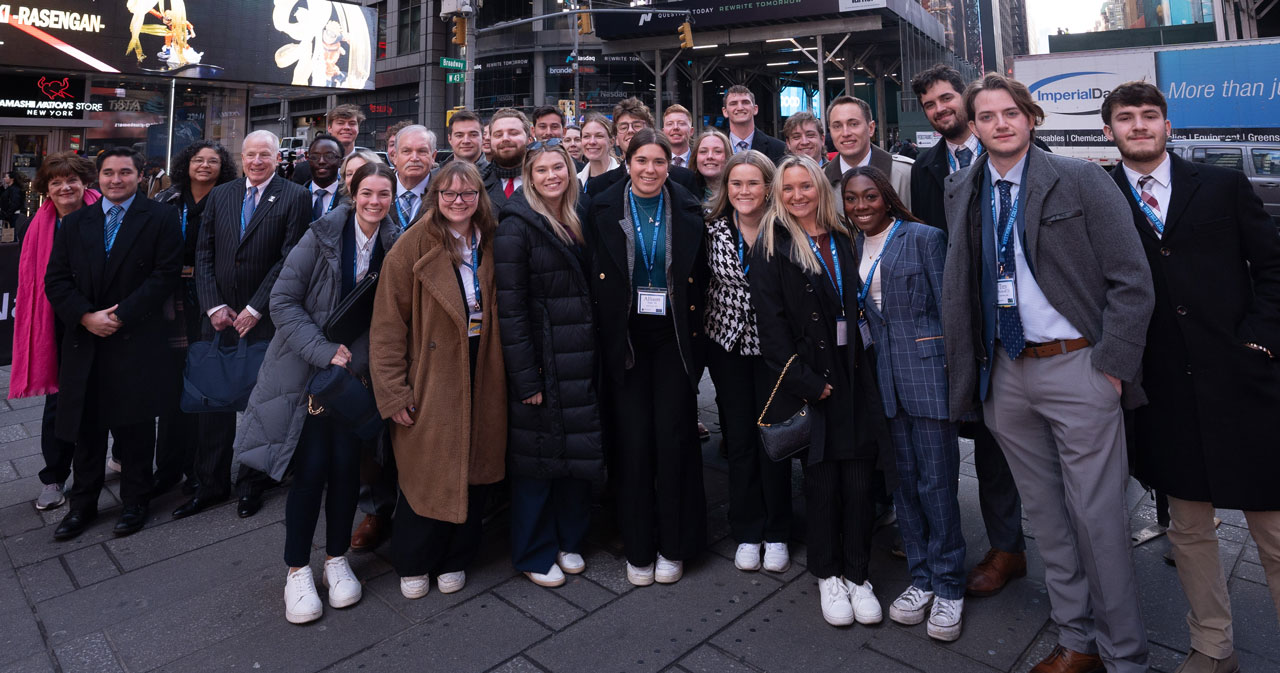 The Blue Blazers Investment Committee poses in front of the Nasdaq sign. Natalie Benton is in front, wearing a long brown coat.