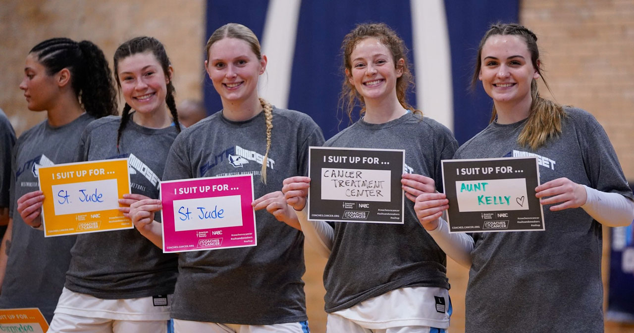 Four smiling women's basketball players hold Coaches vs. Cancer signs on the court of Historic Gymnasium.