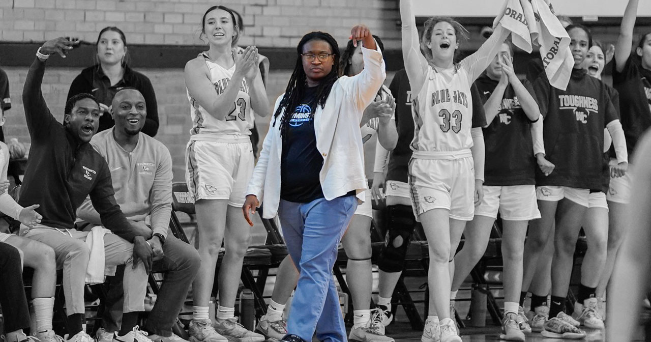 Coach Talisha Washington is pictured in blue pants, white jacket and navy Blue Jays tee, pointing downward and walking on the basketball court, with students and players cheering around her.