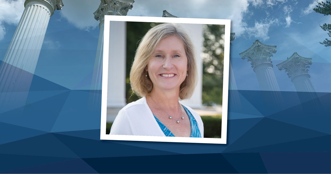 Dr. Mary Majerus is pictured smiling, wearing a white jacket and blue blouse and standing in front of Westminster's Columns. The square photo is placed on top of a blue rectangular background.