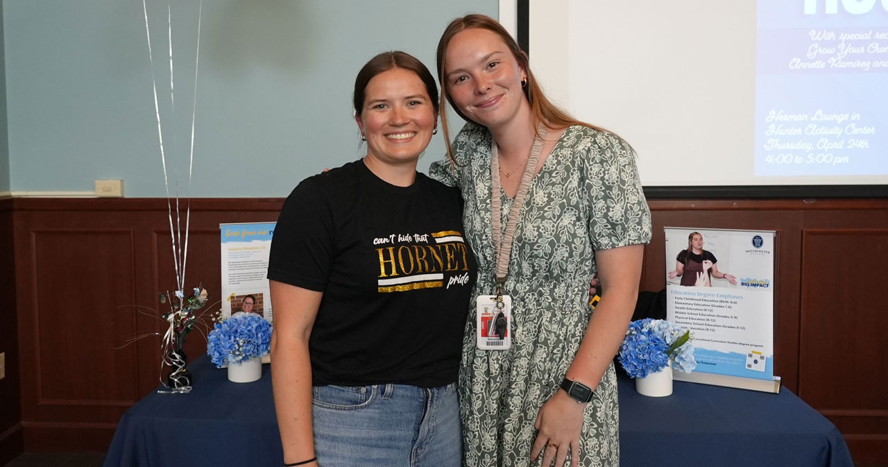 Shelby Kurtz and Madisyn Lenk are picutred, left to right, Kurtz in a black and gold t-shirt and Lenk in a sage green dress, posing in front of an awards table with a blue tablecloth.