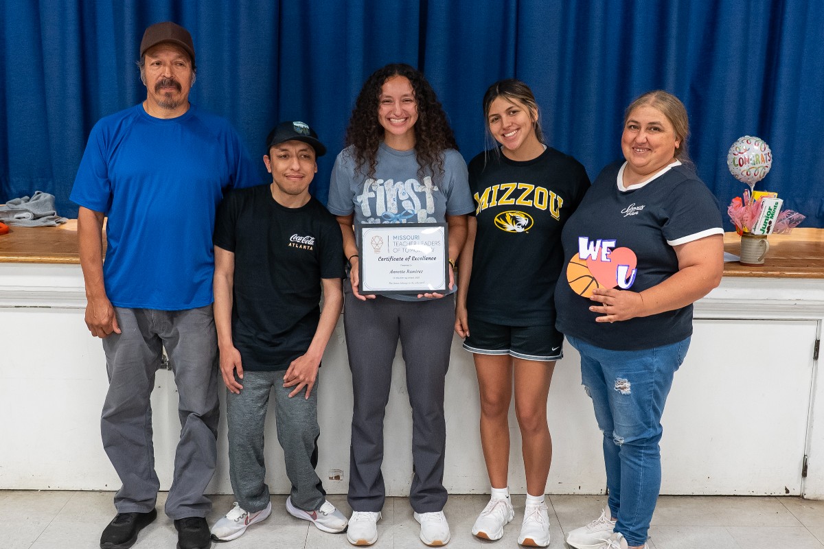 Annette Ramirez is pictured in the middle of her family, standing in front of the Bartley School stage. Her father and brother are to her right in jeans and t-shirts, while her sister and mother are pictured on the left, smiling. Annette is holding an awards certificate.