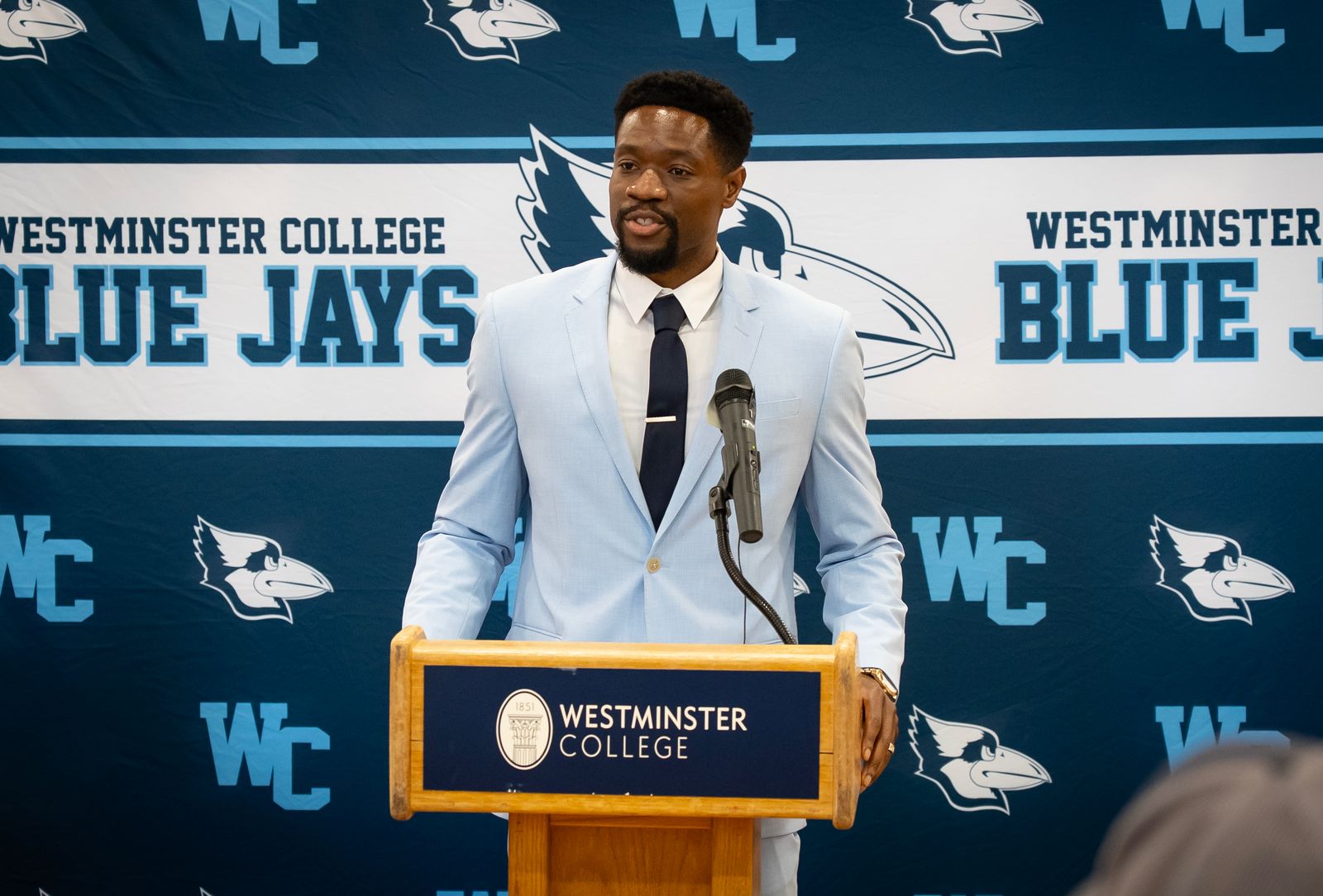 Coach Obi Agomo is pictured wearing a light blue jacket and white shirt, standing at the Westminster College podium in front of a Blue Jays background.