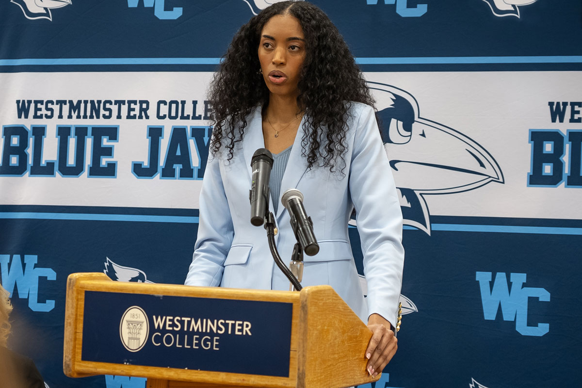 Coach Peyton Whitted is pictured standing at a podium with a Westminster College logo. She is wearing a light blue blazer and is standing in front of a Blue Jays backdrop.