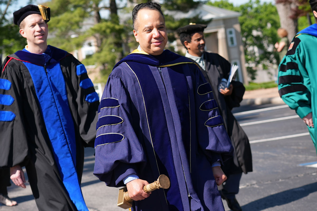 Enrique Salas Durazo is pictured in purple academic regalia walking in a processional outside and holding his Faculty of the Year award in his right hand.