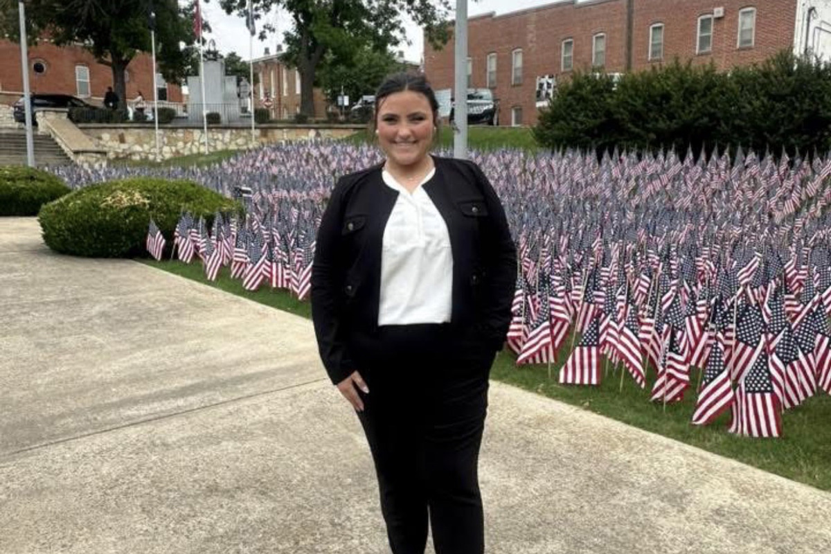 Ailey Fields stands before a lawn covered in tiny American flags. She is wearing a black cardigan and black pants as well as a white blouse. She is smiling.