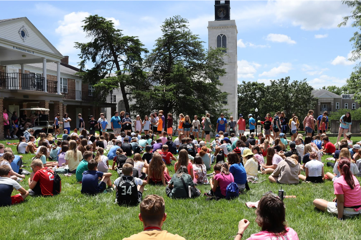 Numerous College for Kids participants sit on the lawn in the Freshman Quadrangle at Westminster, with America's National Churchill Museum in the background. It's a sunny day with blue skies.