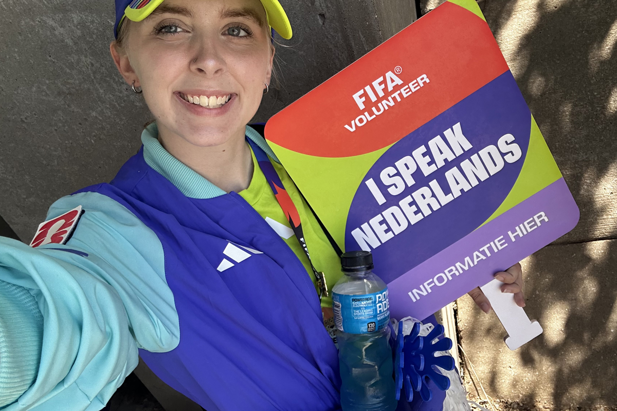 Madison VanDyke is pictured in a light blue shirt and royal blue vest, smiling at the camera a holding an "I speak Nederlands" sign.