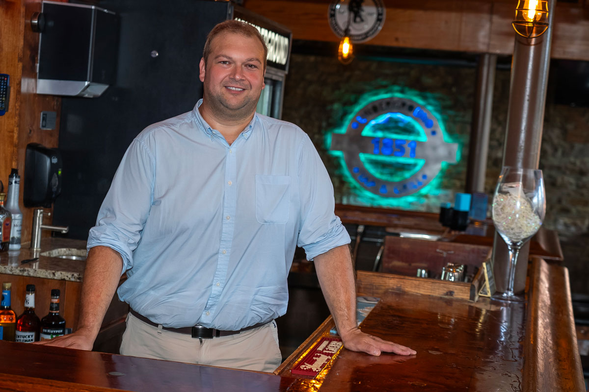 Bryan Hanneke is pictured in a light blue shirt, sleeves rolled up, and khaki pants, standing behind the bar at 1851 Underground with a neon sign behind him.