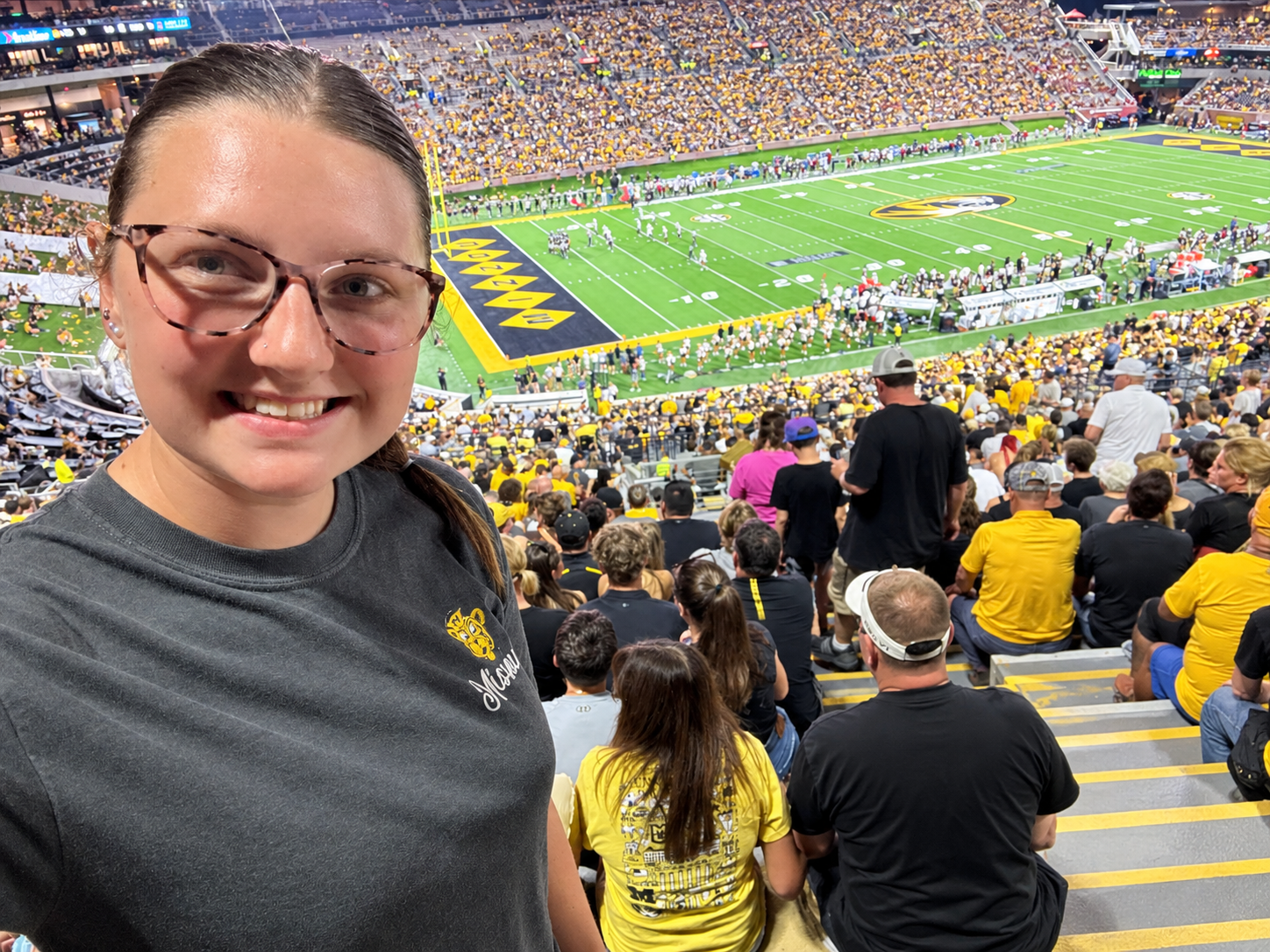Madison Plybon is pictured on the left of the photo with the Mizzou football stadium in the background, seats full. She is wearing a grey t-shirt and glasses.